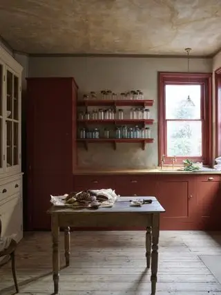 Cuisine rouge brûlé avec cadre de fenêtre peint en rouge, parquet rustique, table carrée et murs texturés.