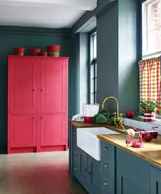 Cuisine peinte en bleu foncé avec armoire en bois autoportante de cuisine rouge vif, parquet en bois clair, évier avec robinet en laiton, demi-rideaux en vichy multicolores