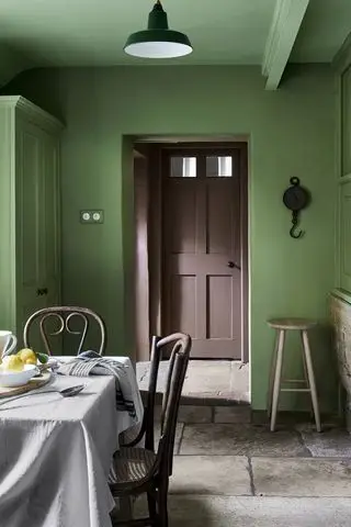 Cuisine de campagne peinte en vert, sol en pierre, table à manger en bois, donnant sur couloir avec porte d'entrée peinte en marron