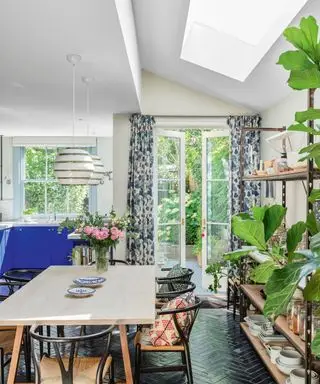 Cuisine salle à manger ouverte, carrelage noir à chevrons, murs peints en blanc, lucarne au plafond, table à manger en bois clair avec chaises en bois noir, rideaux fleuris, grande étagère décorée d'ustensiles de cuisine, deux suspensions blanches au-dessus de la table à manger