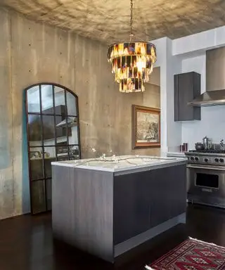 Cuisine moderne avec mur et plafond de style béton, îlot en bois sombre, miroir lambrissé en bois foncé appuyé sur le mur, lustre à plumes au-dessus de l'îlot