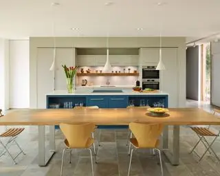 Un exemple d'idées d'éclairage de cuisine moderne montrant une longue table à manger en bois devant un îlot bleu avec des suspensions blanches au-dessus