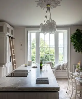 居心地の良い窓側の席を備えたエレガントな白いキッチン、大理石のトップを備えたアイランドキッチン、丸いガラスのペンダントライト、木製の床、白い家具とキャビネット