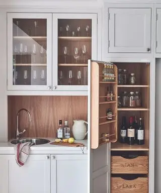 ライトグレーのオーク材のシェーカー スタイルのキャビネット、丸いシンクを備えたバーエリア、ガラス扉付きの吊り戸棚にワイングラスを備えたキッチンの詳細。