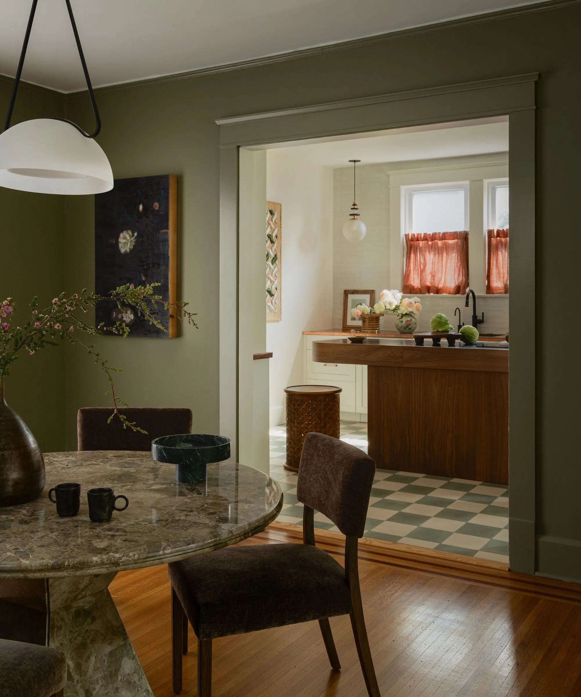 チェッカーボードの床のある居心地の良いキッチン ウッドアイランドのオレンジ色のカフェカーテンとダイニングルームへの開いた出入り口 緑の大理石の丸テーブルのある素朴な緑に塗られた' loading='lazy' title=