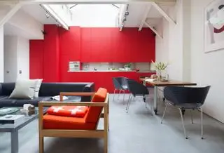 Un agencement de cuisine blanc avec un mur d'accent rouge vif composé d'armoires peintes, avec un canapé gris foncé, des chaises de salle à manger et une table basse.