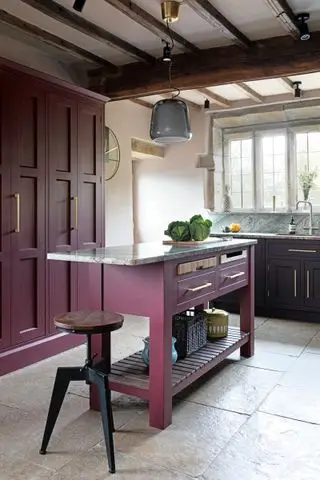 Une cuisine traditionnelle rouge baie avec îlot de cuisine et armoires peints, sol en pierre gris pâle et plafond à colombages.