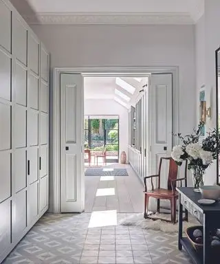 회색과 흰색 타일 바닥으로 마감된 복도, 왼쪽에는 붙박이 찬장이 있고 오른쪽에는 콘솔 테이블과 의자가 있으며 주방과 정원이 보입니다.