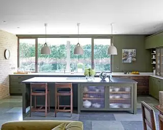 Amplia cocina con isla central, elementos de ladrillo visto, piso de baldosas de color gris oscuro, paredes pintadas de verde, isla con gabinetes de almacenamiento con puertas de vidrio de caña, tres colgantes blancos redondeados que cuelgan sobre la isla, dos taburetes de bar de madera oscura, mesa de comedor de madera oscura en la esquina