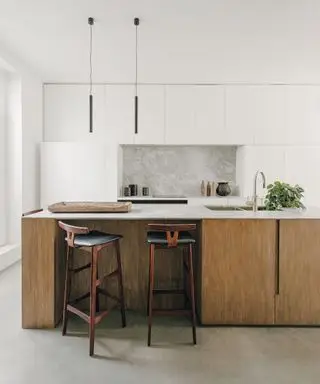 Espacio de cocina moderno con isla de cocina con base y armarios de madera oscura, taburetes de bar de madera oscura y cuero negro, armarios blancos y paredes pintadas de blanco, dos largos colgantes negros colgando sobre la isla