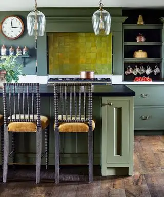 Cocina de estilo tradicional con piso de madera oscura, paredes y gabinetes pintados de verde, pared de azulejos verdes brillantes sobre la estufa, isla de cocina con encimera de granito negro, dos sillas de bar de madera con tapizado de asiento naranja, dos colgantes de vidrio colgando sobre la isla