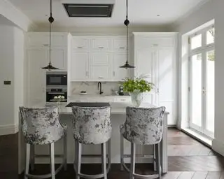 白い食器棚とグレーのベルベットのバースツールのあるアイランドを備えた小さな白いキッチンのアイデア