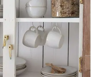 Tasses en céramique blanche suspendues à des crochets sous une étagère d'armoire avec assiettes blanches imbriquées et cuillères en bois en vue