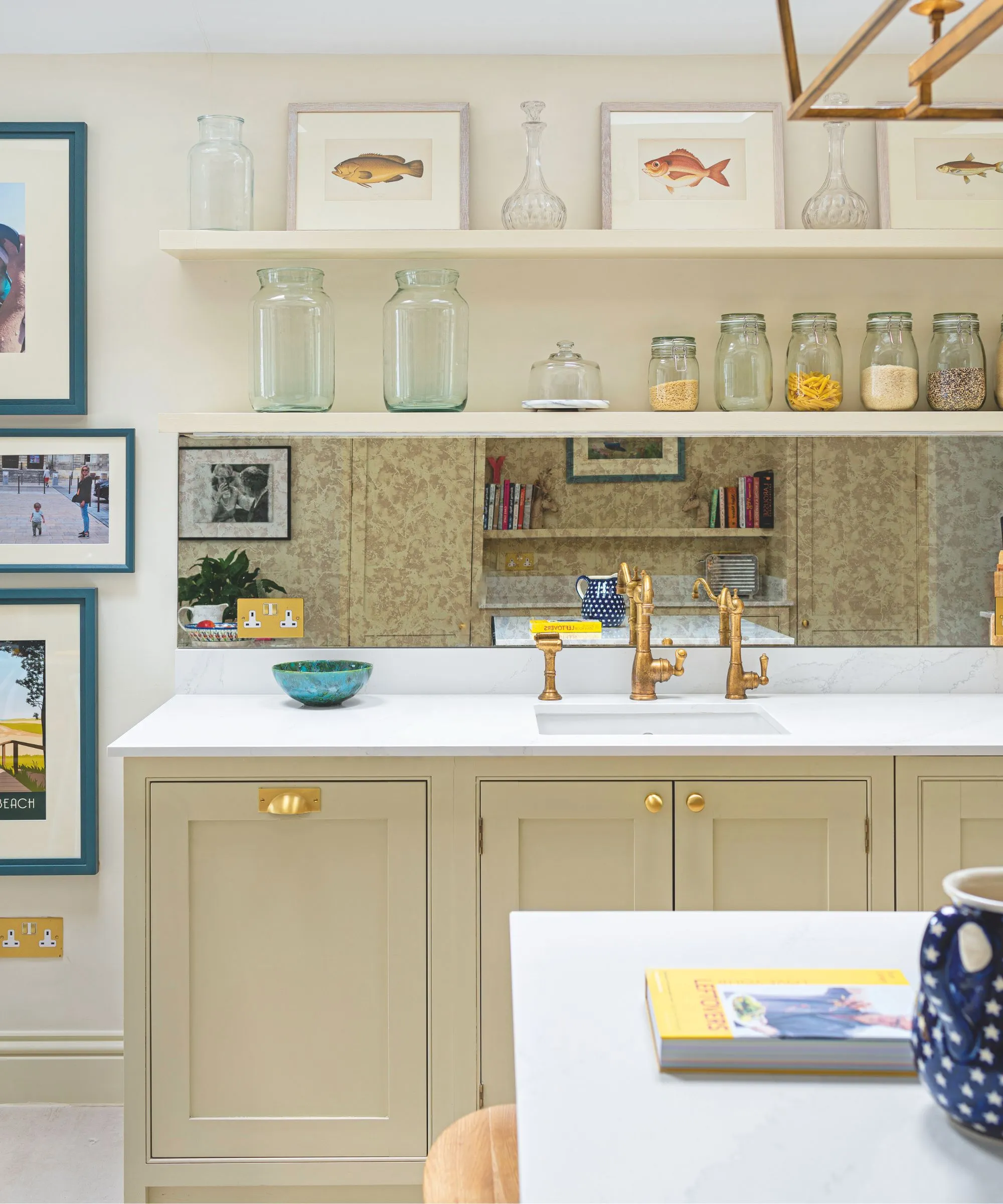 거울 백스플래시 베이지색 캐비닛, 금색 하드웨어, 수도꼭지, 흰색 조리대, 갤러리 벽, 흰색 주방 테이블을 갖춘 크림색 주방입니다.' loading='lazy' title=
