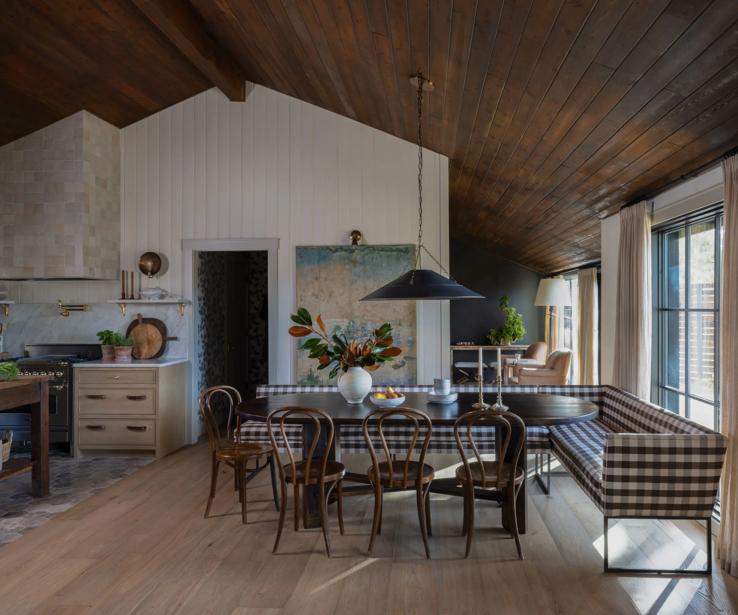 Jídelní kout s tmavým dřevěným oválným stolem obklopeným dřevěnými židlemi a kostkovanou banketou. Na jedné stěně je velký vintage nástěnný závěs a nad stolem visí černé závěsné svítidlo' loading='lazy' title=