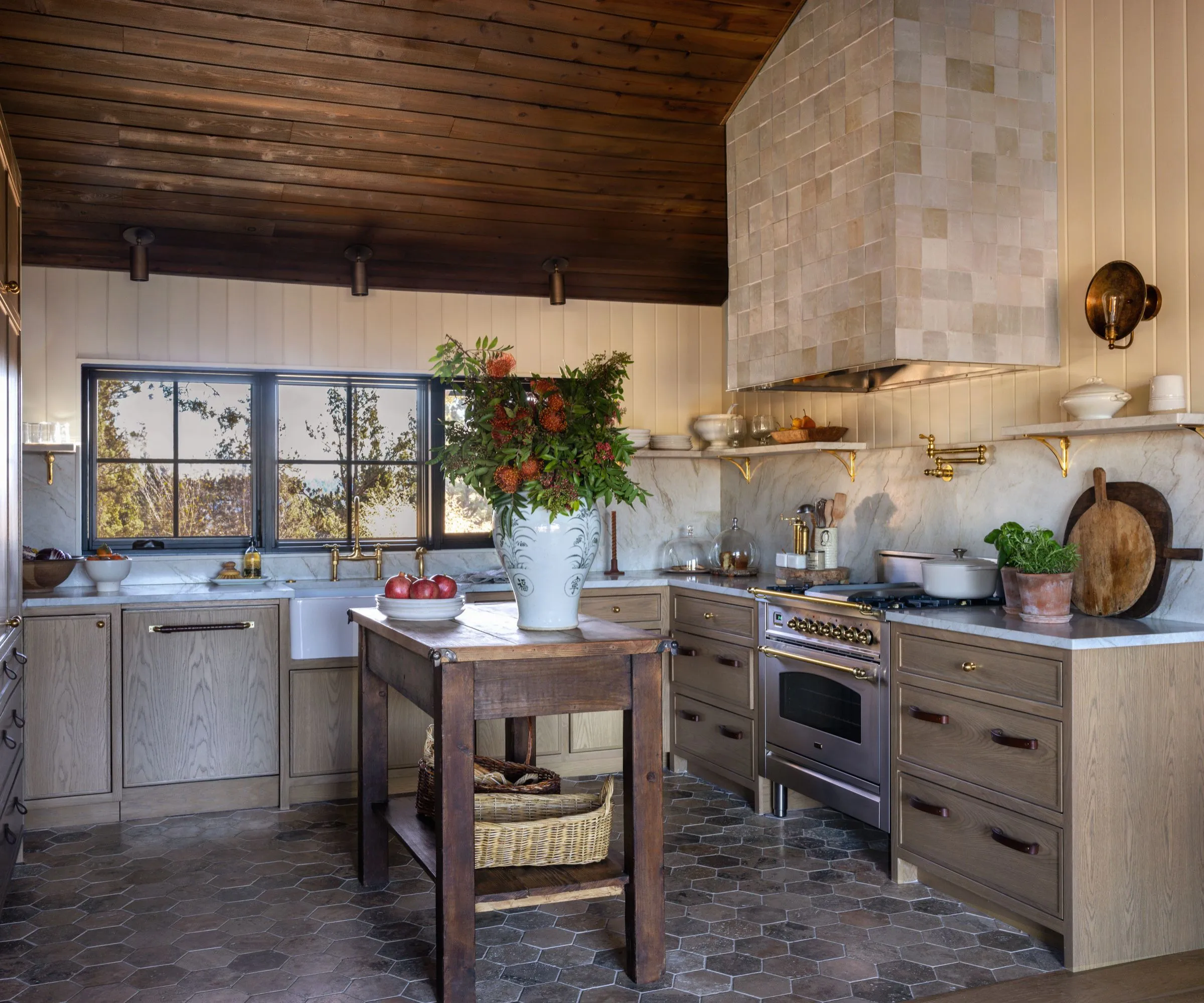 Detailní pohled na pracovní prostor kuchyně s těžkým mramorovým statkovým dřezem, nelakovaným mosazným kováním a integrovanou mramorovou zadní stěnou. Scéna zahrnuje plynový sporák pod minimalistickým omítnutým větracím krytem' loading='lazy' title=