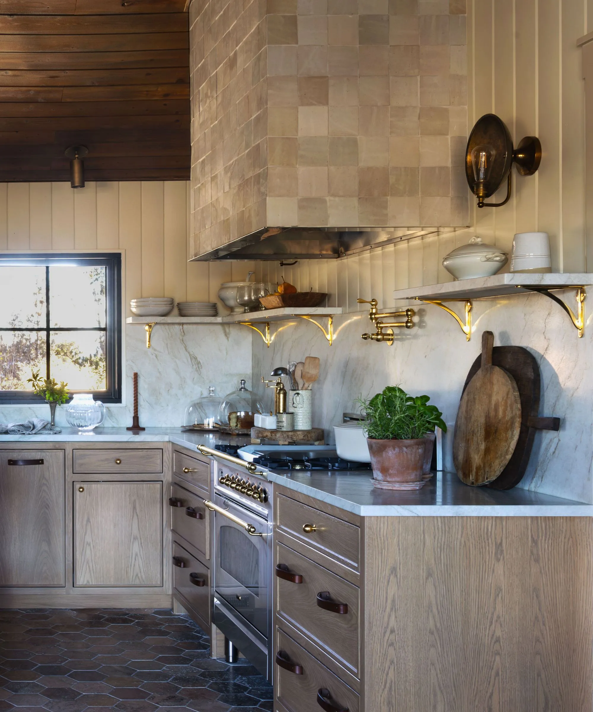 Sluncem zalitý snídaňový kout se zakřivenou vestavěnou banketkou krémové barvy a kulatým dřevěným podstavcem. Nahoře visí mosazné závěsné světlo orámované velkým klenutým oknem a černými skleněnými dveřmi s ocelovým rámem' loading='lazy' title=