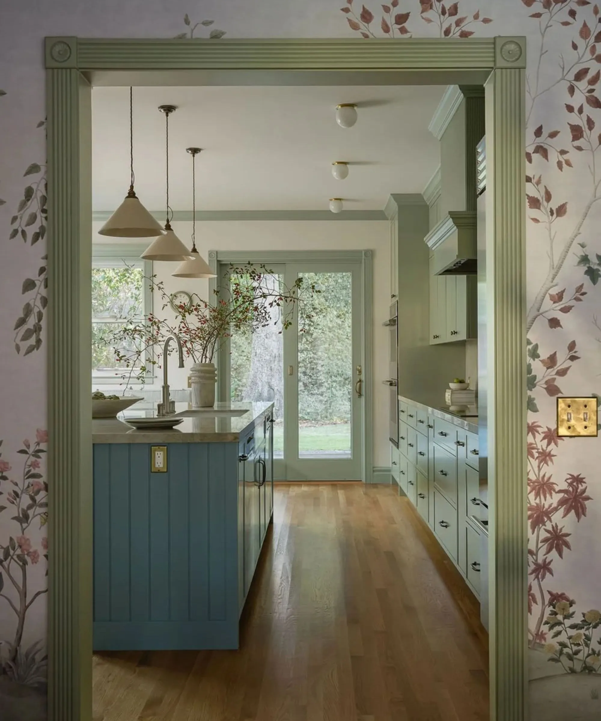 Pohled do kuchyně ukazující kapesní dveře částečně otevřené do spíže. Napravo sahají ke stropu vysoké šalvějové zelené skříně a zlaté vroubkované svícny osvětlují pracovní desku zdobenou velkou zelenou vázou a čerstvými větvemi' loading='lazy' title=