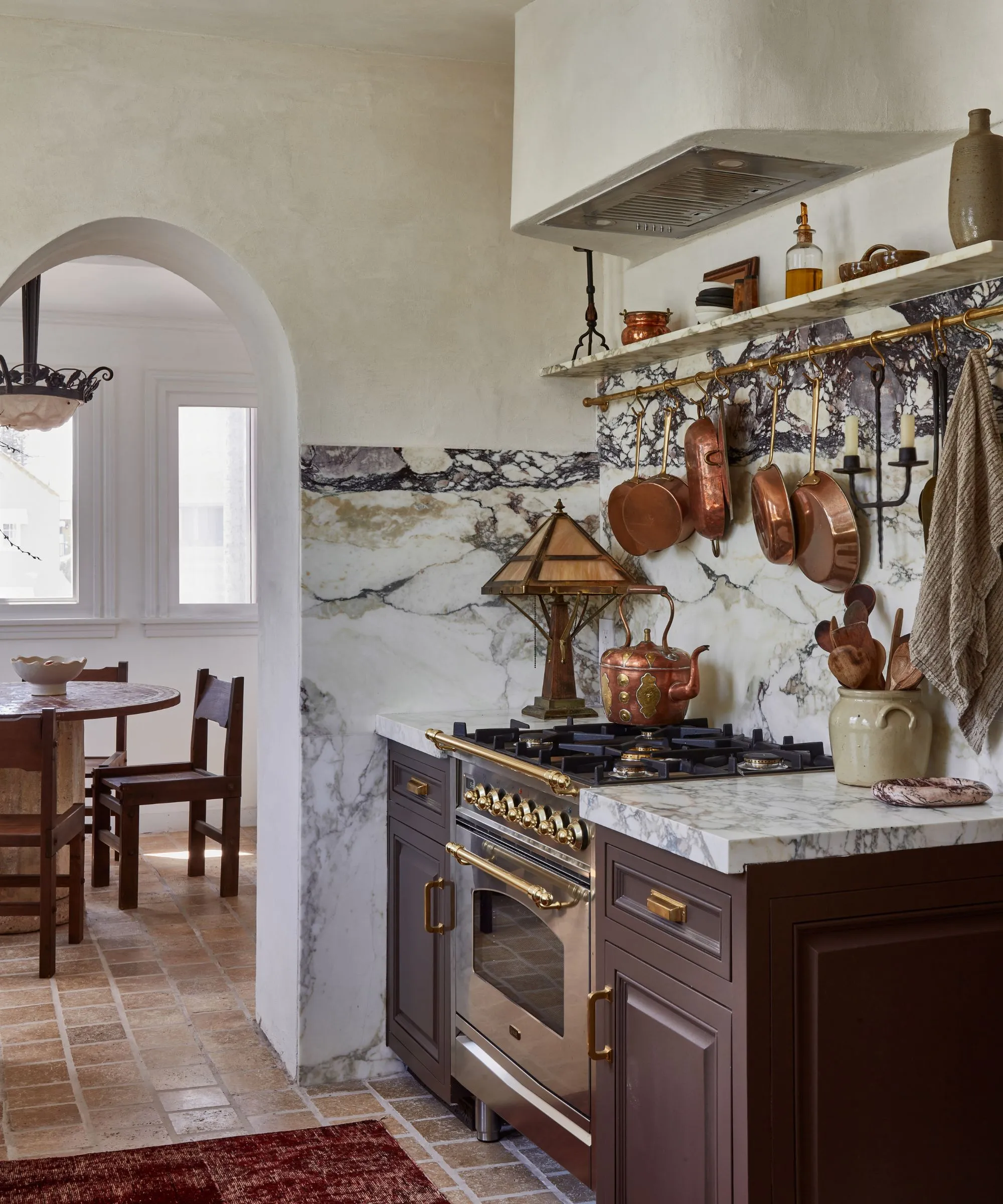 갈색 캐비닛, 대리석 카운터스플래시, 벽에 황동 냄비 걸이, 빈티지 구리 찻주전자를 얹은 레인지 쿠커가 있는 빈티지 주방' loading='lazy' title=