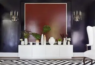 salon violet avec mur rouge derrière le buffet, vases disposés sur le buffet, tapis monochrome, appliques murales de style lustre orné vintage. Fauteuil blanc
