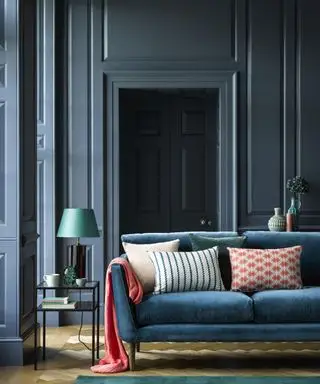 Salón pintado de azul con paneles, sofá azul con coloridos cojines y manta rosa, mesa auxiliar metálica con lámpara de mesa azul, suelo de madera clara