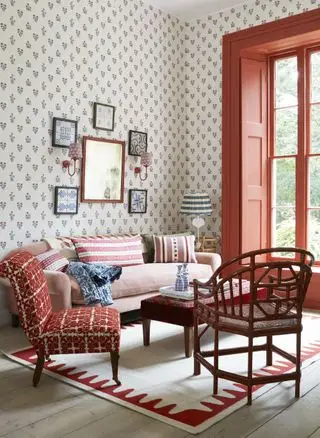 Idea de apartamento con papel tapiz con letras pequeñas, carpintería pintada de naranja alrededor de la ventana, sofá rosa, sillas y mesa de café rojas, alfombra color crema y roja, tarimas encaladas, obras de arte, apliques y lámpara de mesa.