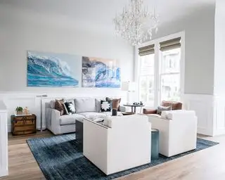 Salón costero con dos obras de arte en la pared, pared pintada de gris claro, gran lámpara de araña de cristal, alfombra azul sobre suelo de madera, dos sillones color crema frente a un sofá gris con mesa de centro gris oscuro