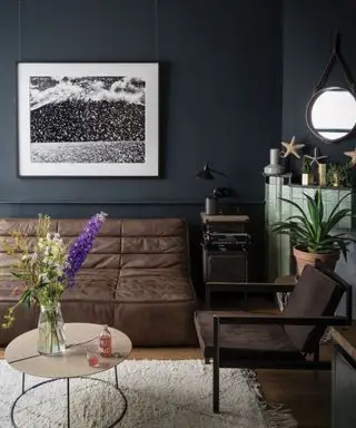 Sala de estar pintada de gris oscuro por Farrow & Ball, sofá de cuero marrón, chimenea con azulejos verdes, piso de madera con alfombra color crema, techo pintado de blanco, obras de arte en blanco y negro en la pared, accesorios costeros y diseño de espejo marítimo sobre la chimenea