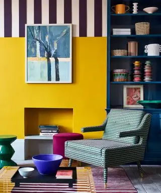 Esquina colorida de una sala de estar, pared pintada de amarillo, chimenea, sillón tapizado, diseño pintado con rayas en la pared superior, estantería pintada de azul, obras de arte en las paredes, libros y adornos en la estantería