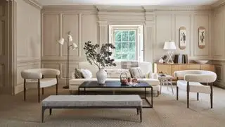 Beige gestrichenes Wohnzimmer mit Täfelung rund um den Raum, geräumiger Sitzbereich mit cremefarbenem Sofa und zwei Sesseln aus Bouclé-Stoff, schwarzem rechteckigem Couchtisch und grauem Fußhocker, weißer Metall-Stehlampe, Fenster mit Fensterladen, hellem Holz-Sideboard mit weißer Tischlampe und Accessoires