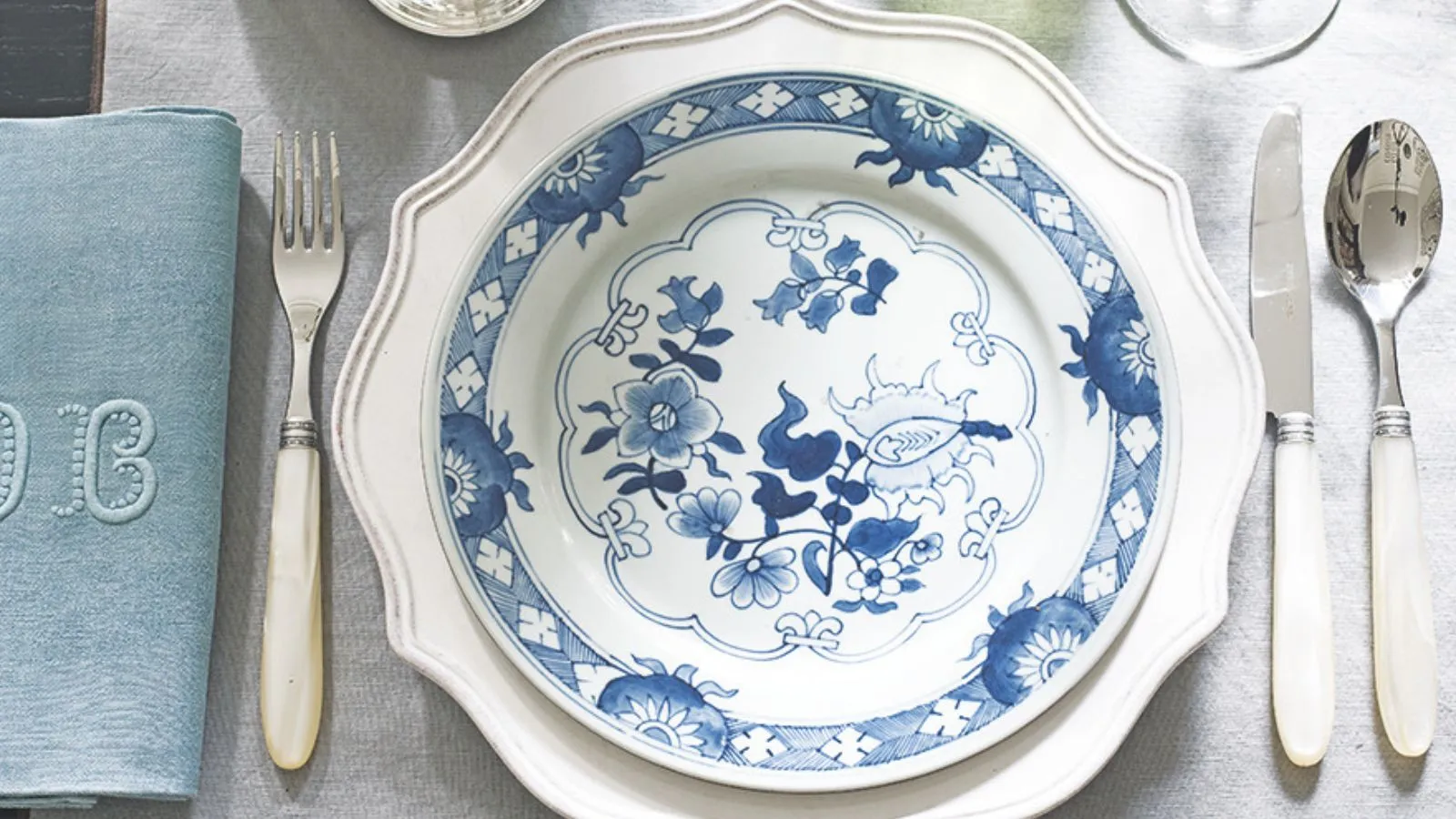 Bílý stůl s bledě modrým bílým příborem ubrousku a bílou nabíjecí deskou s modrým a bílým vzorovaným nádobí' loading='lazy' title=