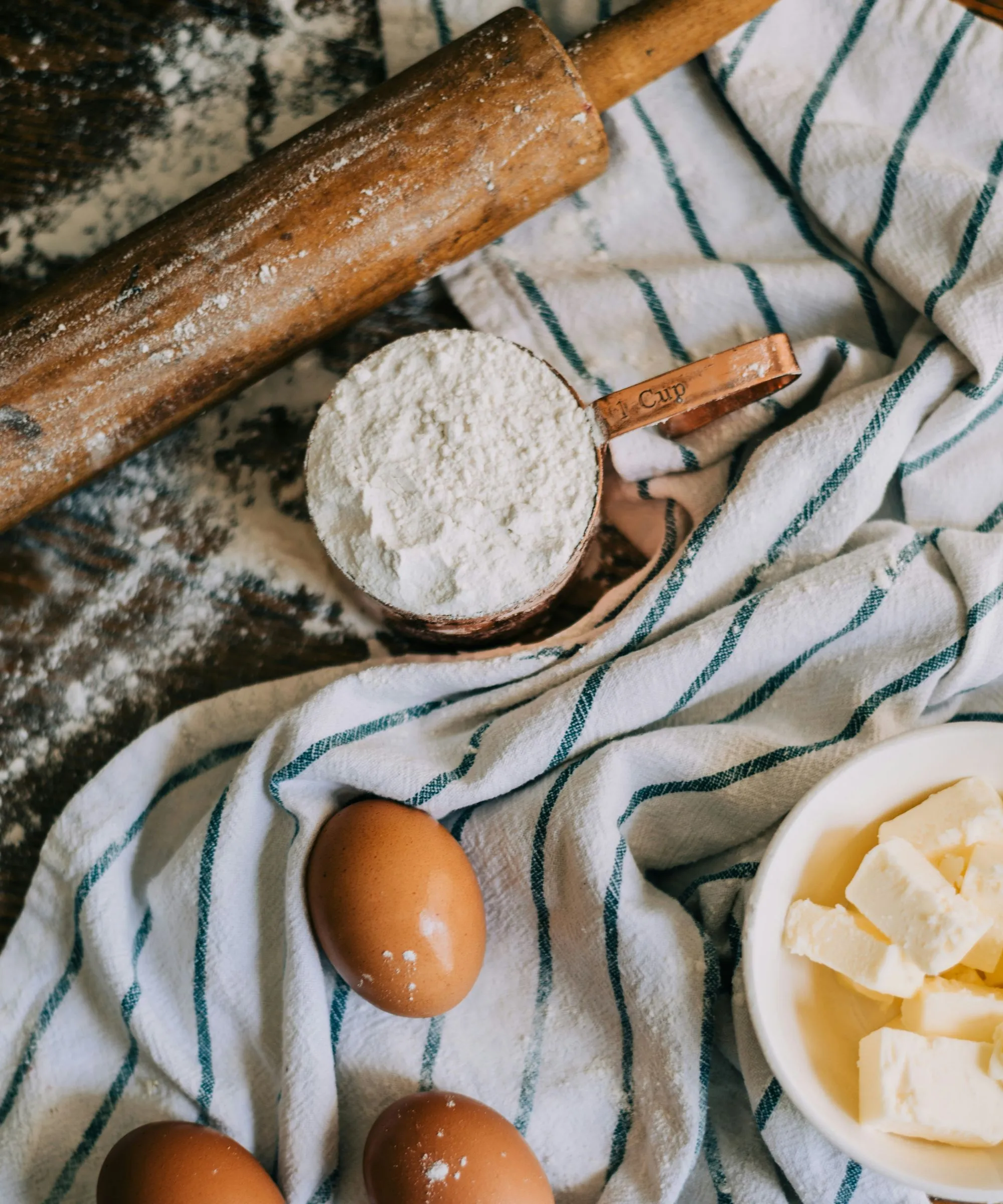 나무 롤링 핀 옆에 파란색과 흰색 줄무늬 차 타월 위에 계란 버터와 밀가루' loading='lazy' title=