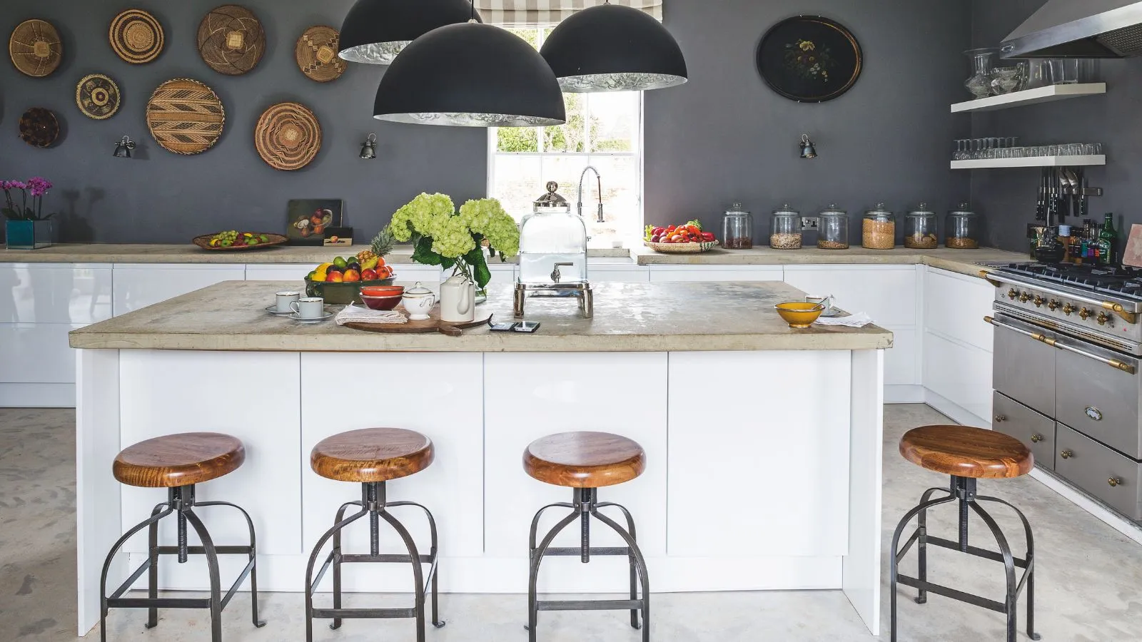 大理石のカウンタートップ、木製と金属製のバースツール、丸いステートメント ペンダント ランプ、壁に装飾プレートを備えた白いキッチン アイランドを備えたグレーのキッチン' loading='lazy' title=