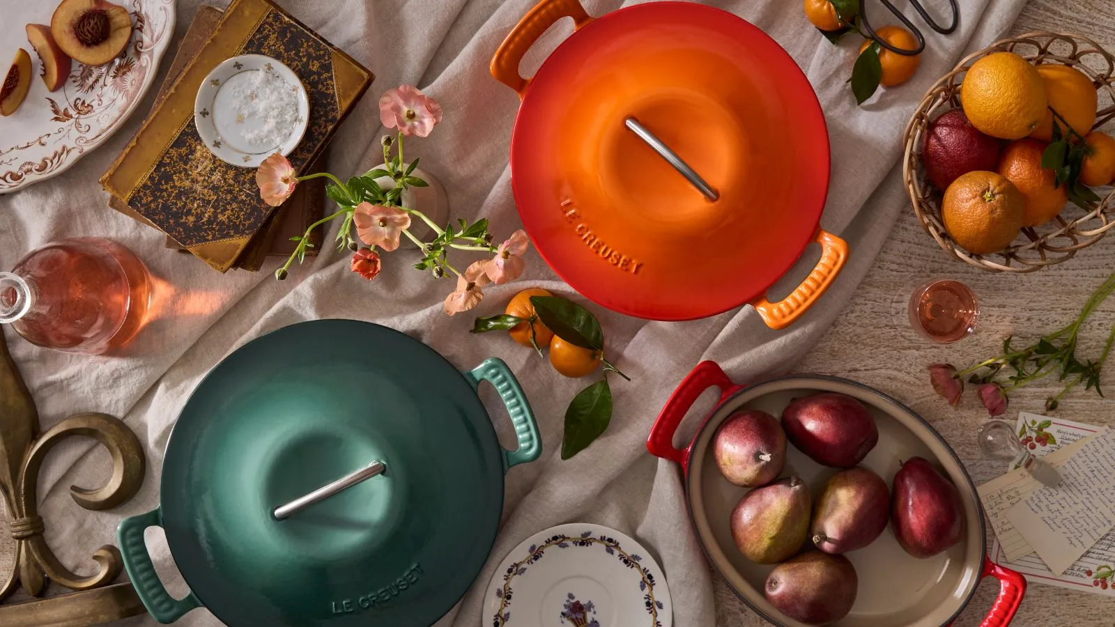 Zelená a oranžová holandská trouba z nové kolekce Le Creuset Modern Heritage Sbírka shora obklopená čerstvým ovocem a květinami' loading='lazy' title=