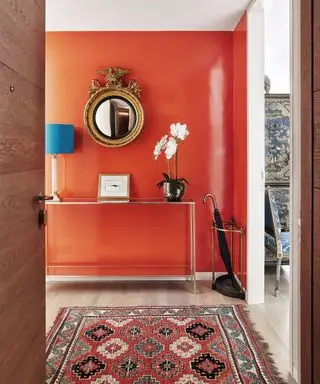 Couloir avec mur d'accent rouge-orange, table console en verre et métal et tapis orange à motifs géométriques sur parquet.