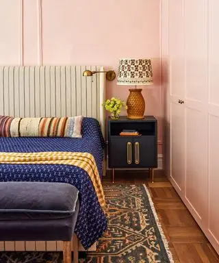 couleurs qui vont avec le rose clair, chambre aux murs rose clair, lit rembourré à rayures, couverture bleu indigo, table de nuit bleu marine, pouf bleu marine, tapis vintage, parquet, applique murale, abat-jour à motifs sur lampe de table, oreiller de lit à rayures, couverture à carreaux curcuma, armoires