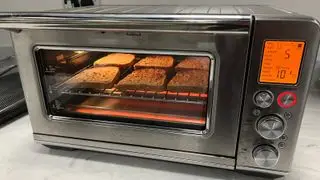 Breville the Smart Oven Air Fryer Toaster Ovn som lager toast på tekst. Den lyses nedenfra av varmeelementet og skjermen på venstre side lyser oransje, med funksjonene og tidene oppført.