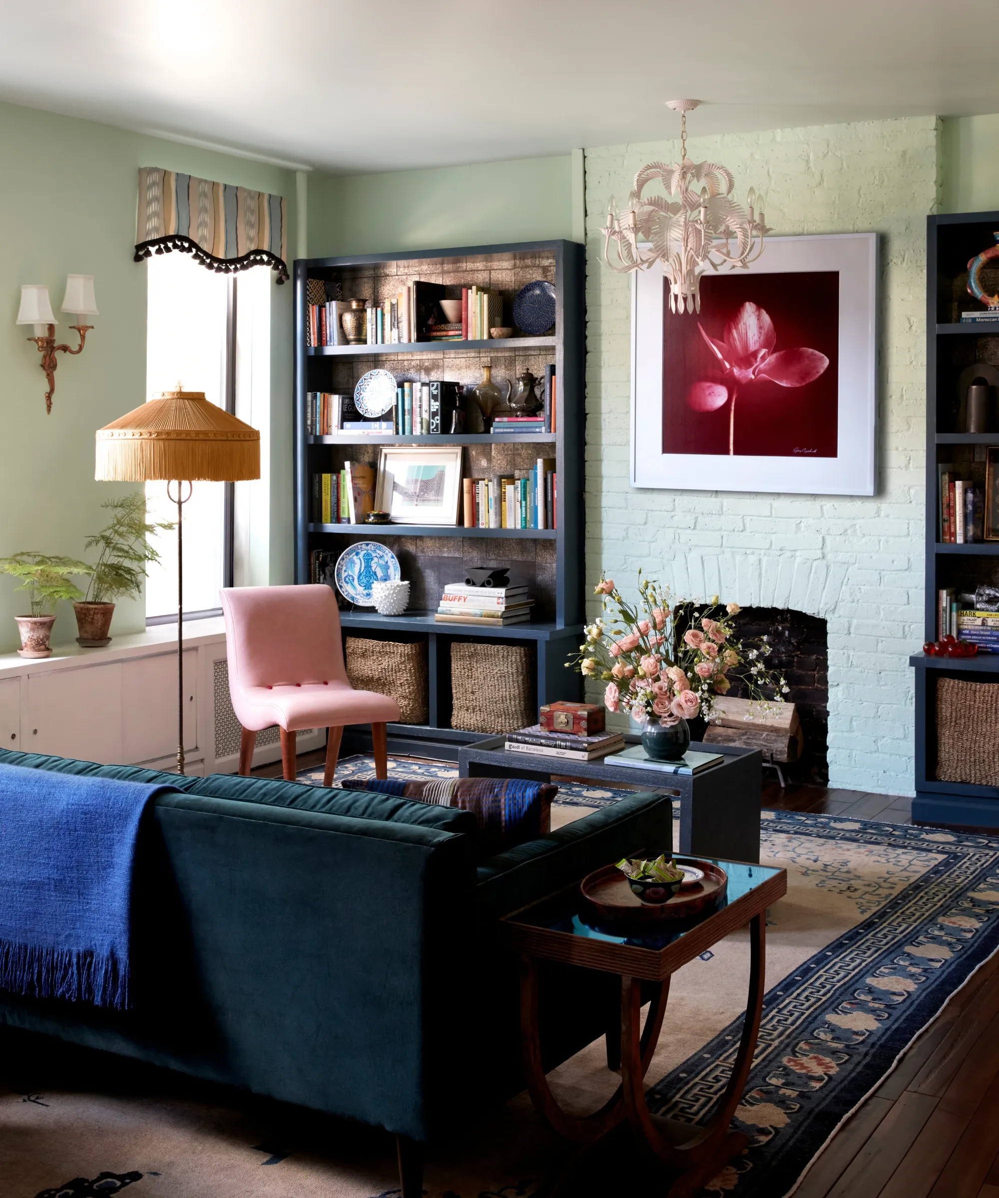 Kolorowy salon z jasnozielonymi ścianami, ciemnoniebieską sofą, abażurami z falbankami, wzorzystym dywanikiem i półką pełną książek i różnych przedmiotów.' loading='lazy' title=