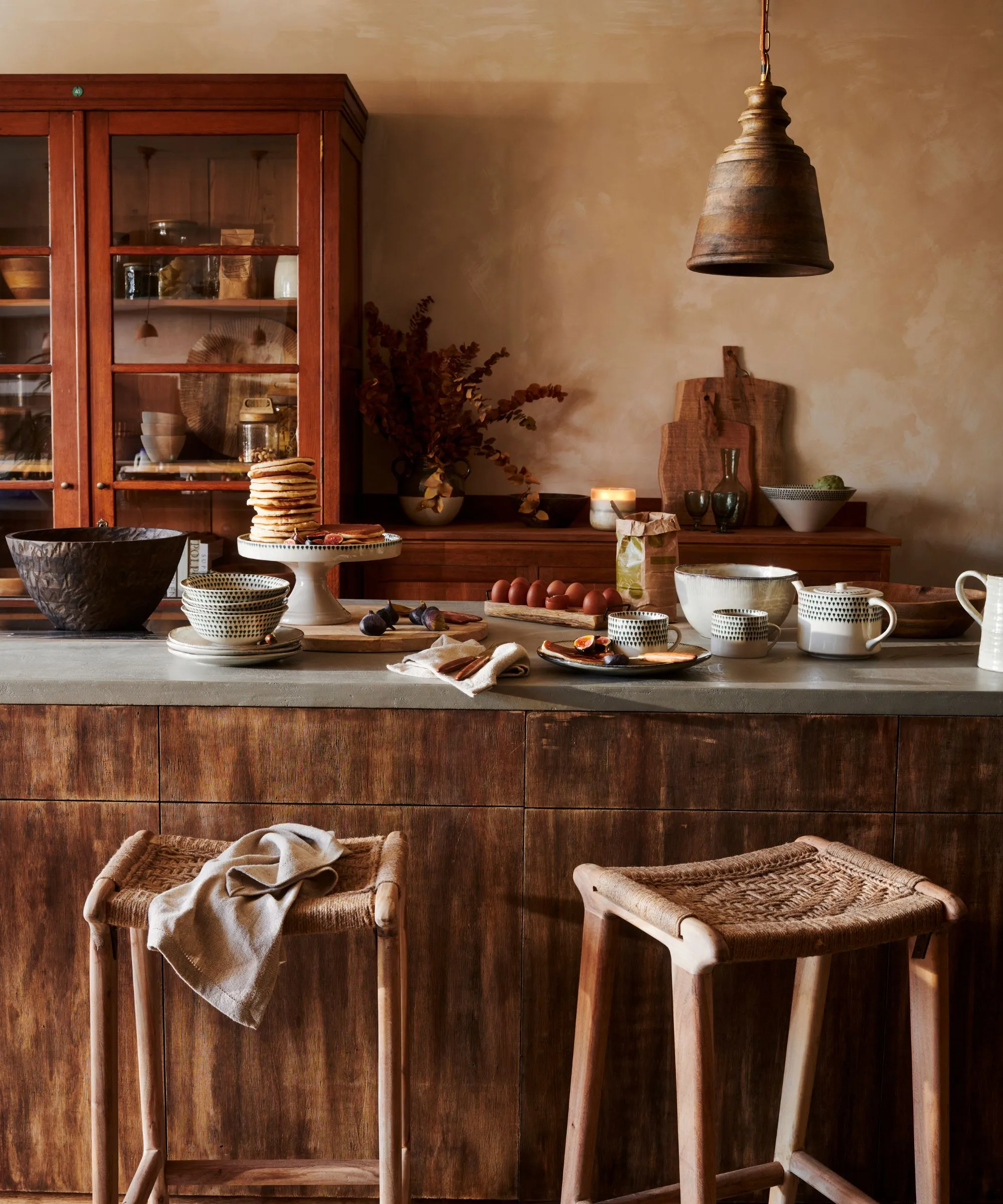 Una cocina en tonos tierra con isla de madera y despensa pintada de rojo. Los panqueques se apilan en un puesto de pasteles en la isla. Se acercaron dos taburetes de ratán.' loading='lazy' title=