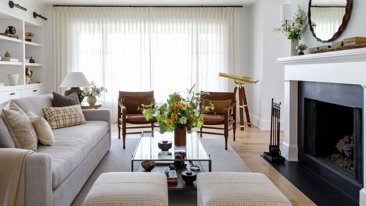 סלון לבן בהיר עם אח גדול וילונות שקופים מהרצפה עד התקרה מובנים מדפי ספרים לבנים ורצפות עץ' loading='lazy' title=