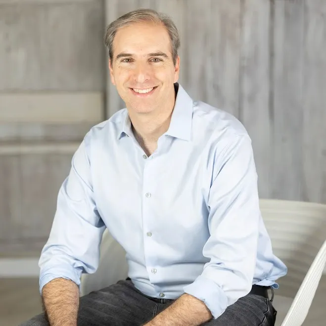 zdjęcie profesjonalnego organizatora Bena Soreffa siedzącego na białym krześle na drewnianym tle, ubranego w jasnoniebieską koszulę z długim rękawem i czarne dżinsy' loading='lazy' title=