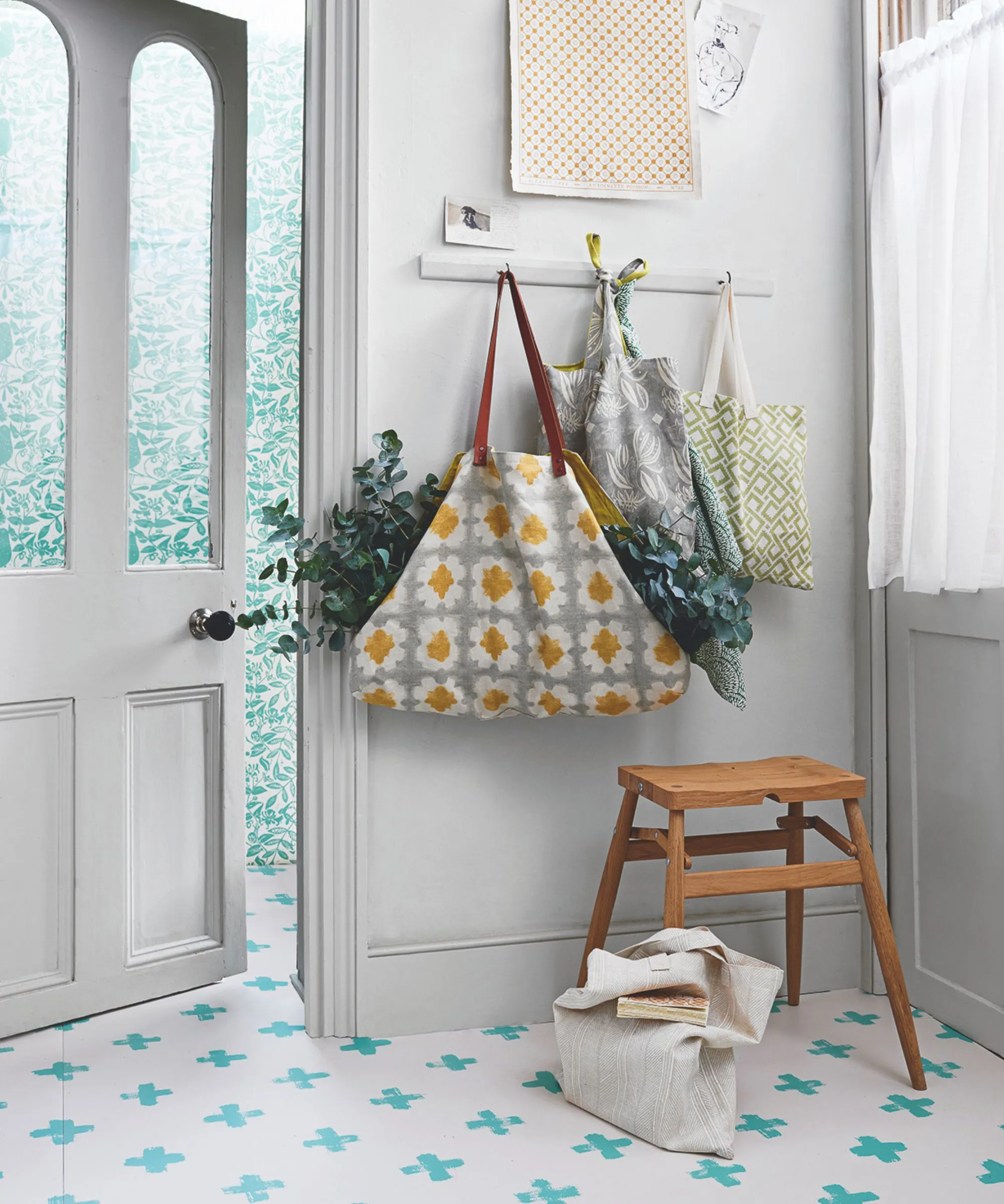 Biały korytarz z niebiesko-białą podłogą wyłożoną kafelkami, białymi szklanymi drzwiami i wieszakami ściennymi z torbami wypełnionymi roślinami. Pod nimi znajduje się drewniany stołek, a po prawej stronie okno z przezroczystą zasłoną.' loading='lazy' title=