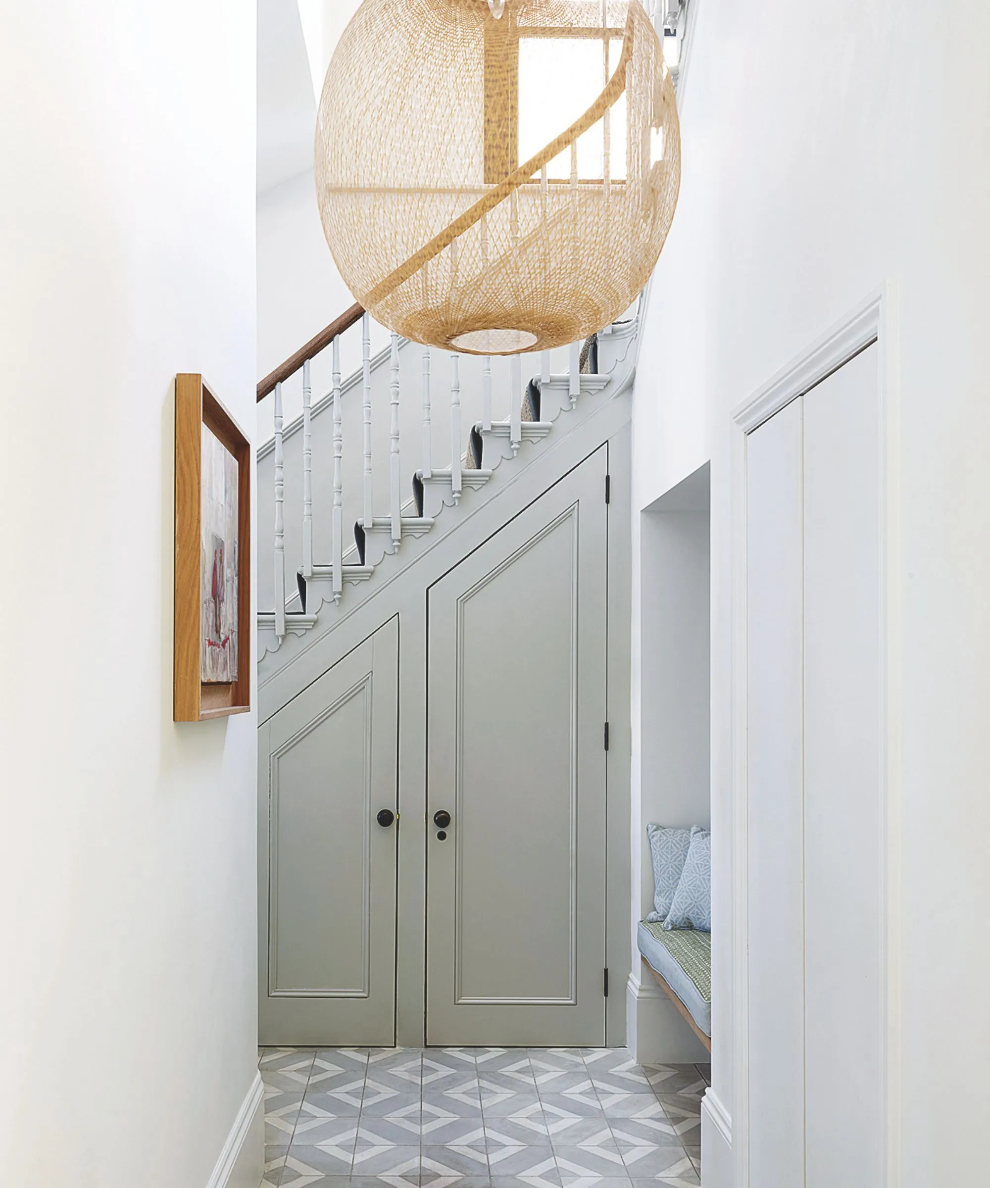 Biały przedpokój z podłogą wyłożoną kafelkami, wbudowana rattanowa ławka z grafiką w drewnianej ramie na ścianie i duża rattanowa lampa wisząca nad nią.' loading='lazy' title=