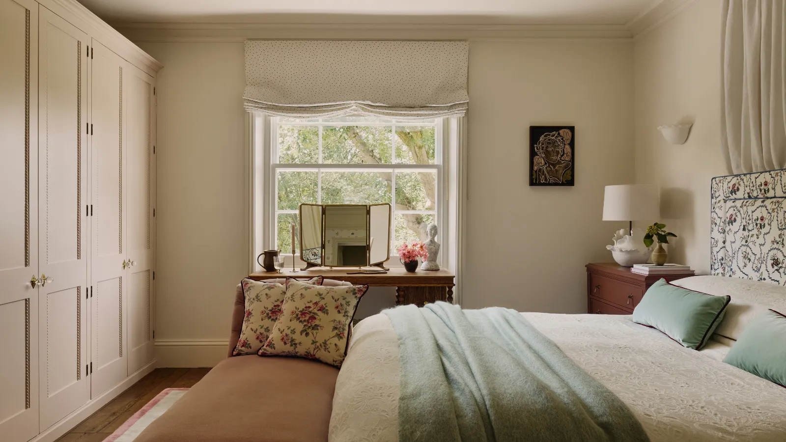 una camera da letto con pareti dai colori neutri una parete di armadi una grande finestra un letto con lenzuola color crema una coperta verde acqua e cuscini verde acqua e una panca ai piedi del letto con due cuscini floreali' loading='lazy' title=