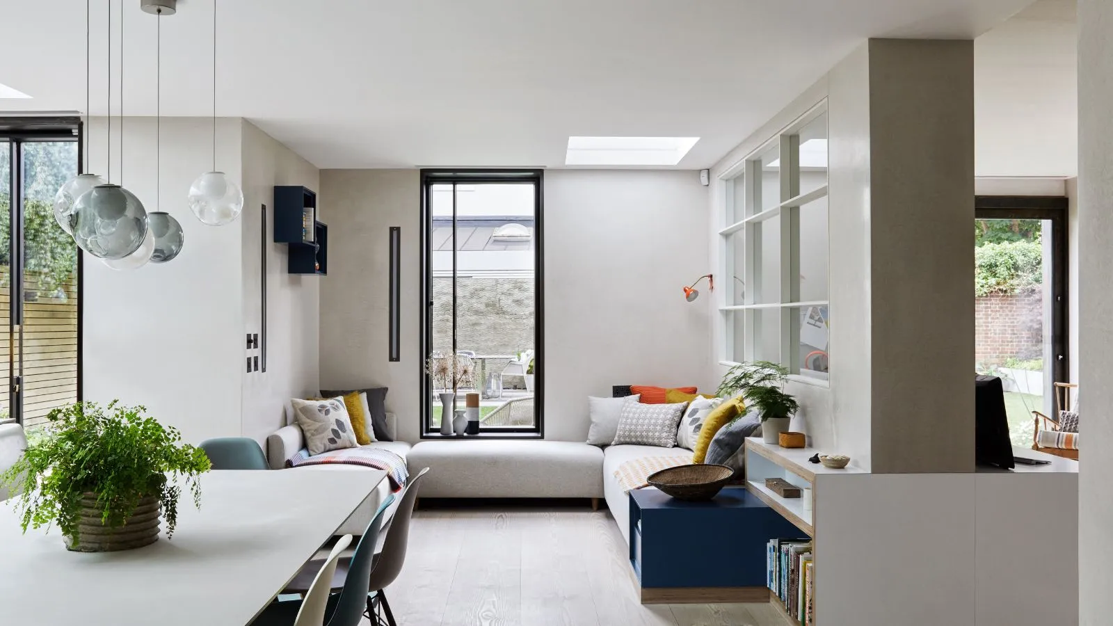 Ein heller Sitzbereich mit natürlichen Licht aus großen Fenstern mit hellem Holzboden, ein cremefarbenes Ecksofa und ein cremefarbener Tisch mit hängenden Glasheizenleuchten, die darüber hängen' loading='lazy' title=