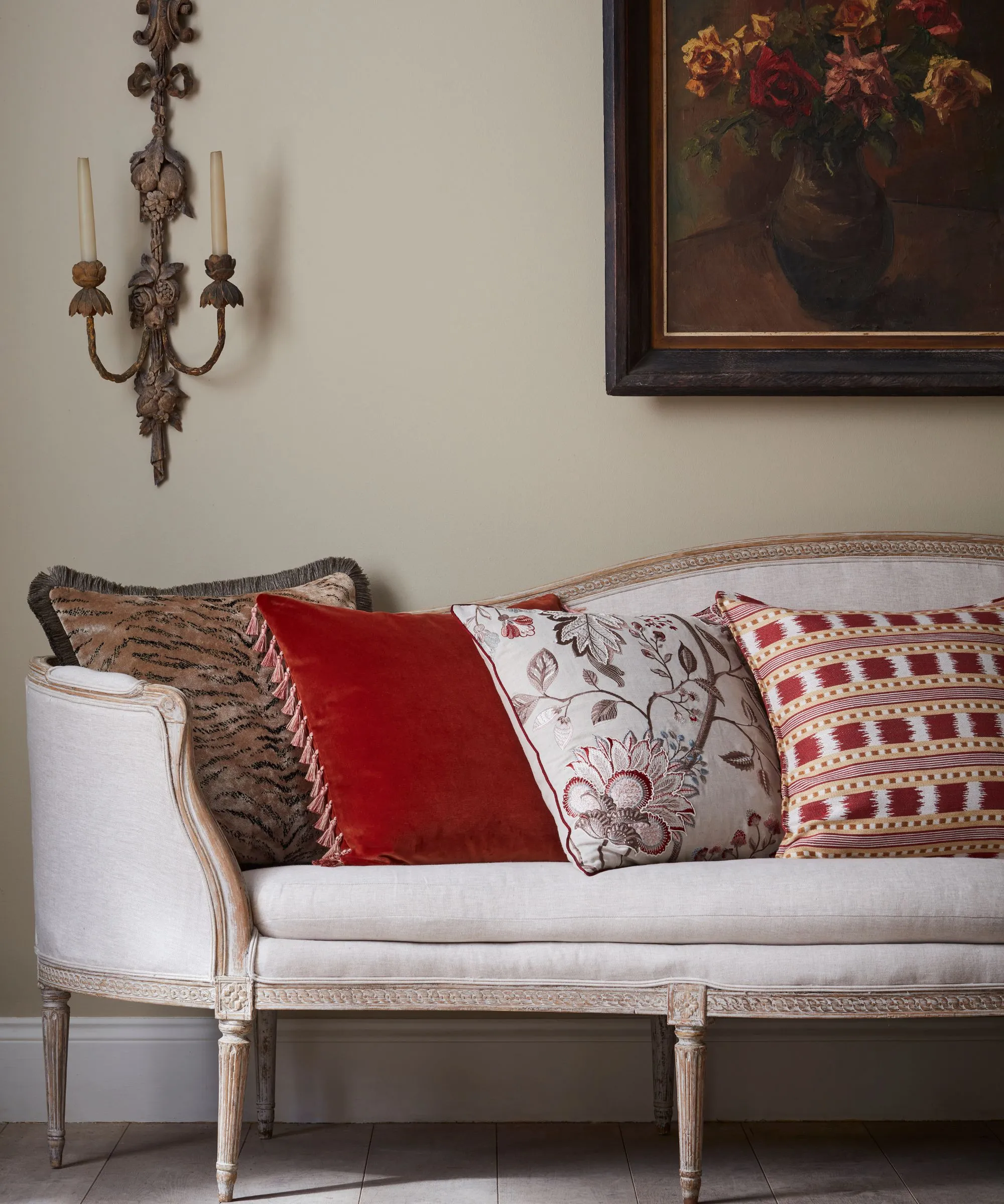 Chaise longue tapizada en blanco de aspecto vintage con cuatro cojines en diferentes estampados y diseños en blanco, rojo, marrón y gris topo. Hay un candelabro ornamentado montado en la pared con velas delgadas y arte enmarcado en la pared con flores otoñales.' loading='lazy' title=