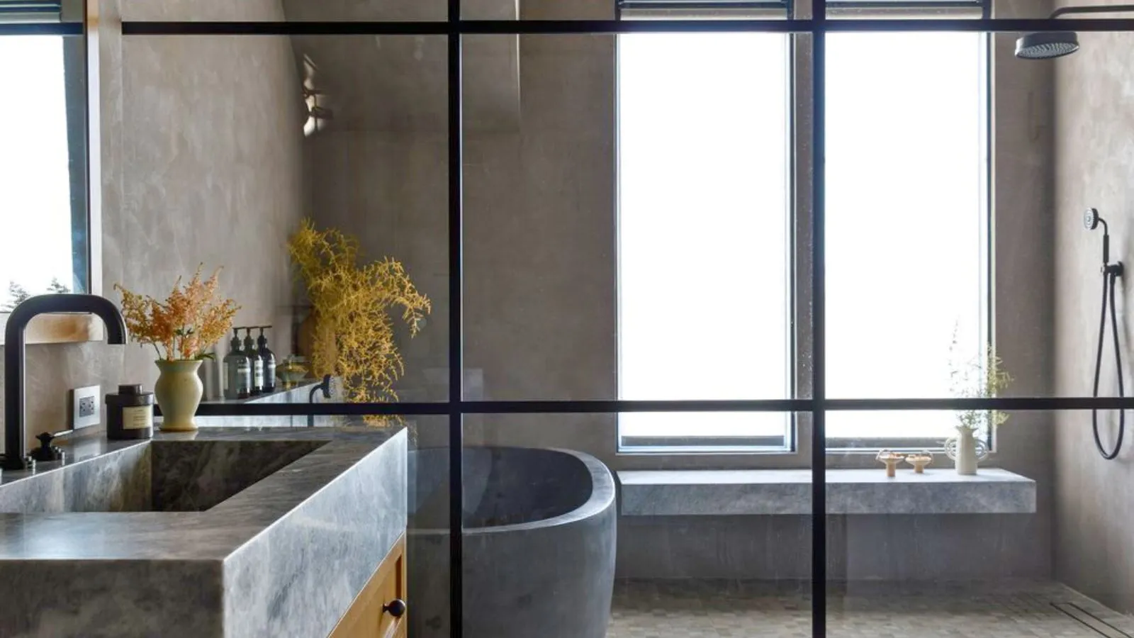 Sklenená sprcha Crittal v neutrálnej kamennej kúpeľni. Vidno tu sprchové sedadlo a veľké matné okno, ako aj sivú kamennú vaňu a kamenné umývadlo' loading='lazy' title=