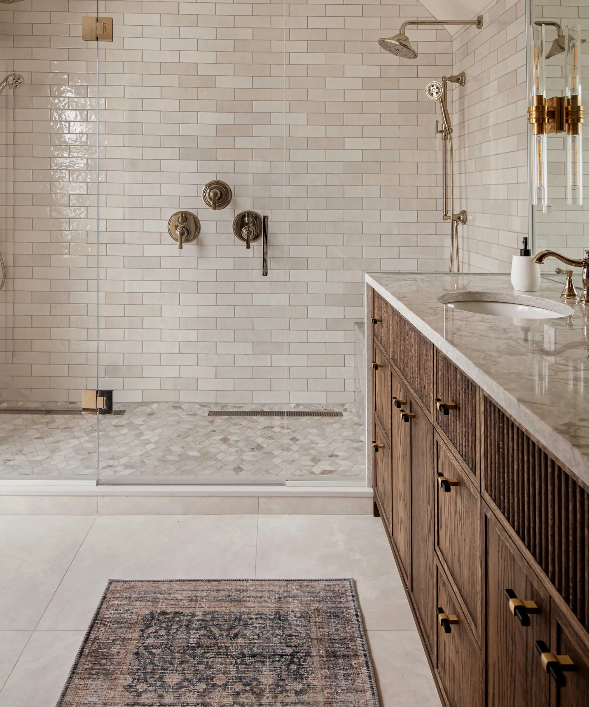 kúpeľňa so sivobielymi kachľovými stenami v sprche, béžová mramorová doska pre skrinky z tmavého dreva pod ňou a perzský bežecký koberec na podlahe' loading='lazy' title=