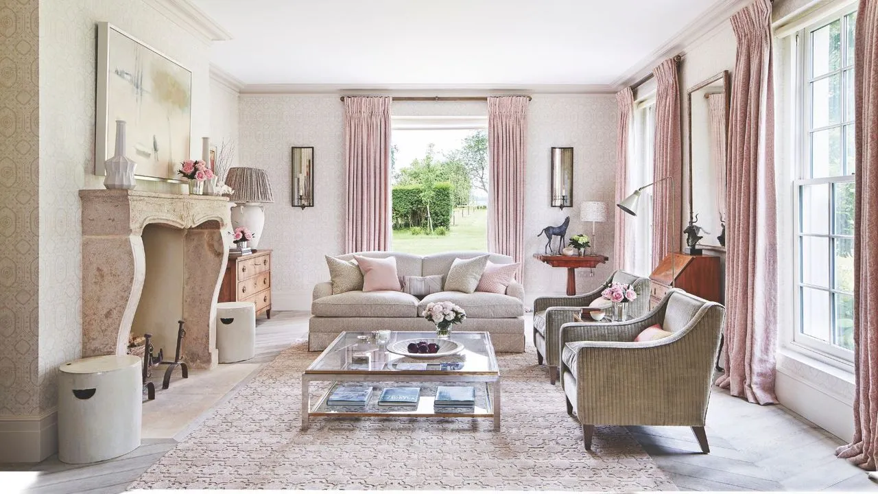 Różowy salon z różowymi zasłonami, różowym wzorzystym dywanikiem, szklanym stolikiem kawowym, neutralnymi kanapami i dużym kamiennym kominkiem. Ściany są białe, a przez okna wzdłuż ścian widać zielony ogród.' loading='lazy' title=