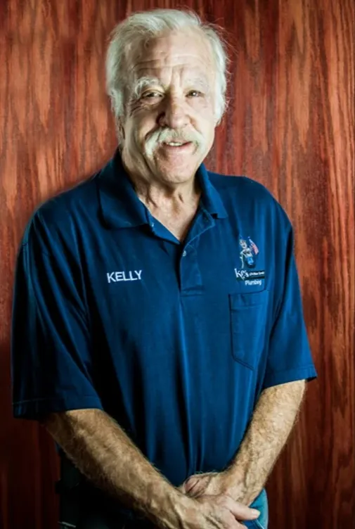 Kelly Russum w niebieskiej koszulce polo przed brązowym drewnianym tłem.' loading='lazy' title=